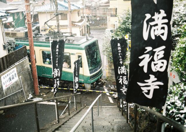 2026年2月8日の雪窓の外を布団を被りながら這いつくばって見に行くと、雪が降っていた。住んでいる家は築60年を超えるため、保温という概念がなく、外よりも寒い。冷蔵庫のようなものだ。そういう意味ではある意味、保温されている。信じられないほど寒い中、気合いで起きて、即効着替えて、フィルムカメラ片手に近所を散策。雪が降ると、やっぱりちょっとワクワクしてしまう。カメラを操作しやすいように、ミトン型で指先だけが露出しているタイプの手袋をしていったため、指先は壊死するのではないかというほど血色悪く、感覚は失われている。一刻も早く、こたつに入りたいという思いと、もう少し写真を撮りたいという思いが交差する。14分に1本の江ノ電をポイントポイントで待つわけだが、静止してしまうと内臓が震え始めてしまうので、意味もなく動き続ける。江ノ電が来た瞬間、壊死寸前の人差し指でシャッターを押す。カシャン！雪の降る音が聞こえそうなほどの静寂、ではなく、江ノ電の余計にうるさいエンジン音と、線路との摩擦音にかき消されるようにシャッター音が反響する。来年も降るかなあ。#snow #enoden #満福寺 #江ノ電 #鎌倉